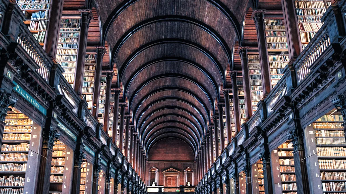 Sala de leitura de uma biblioteca antiga, com estantes altas de livros e uma abóbada de madeira por cima.
