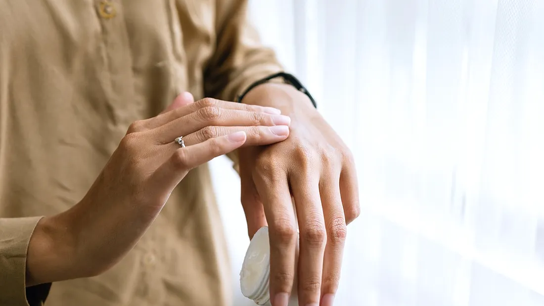 Mãos a espalhar creme no dorso da mão, junto a uma janela com luz de dia.