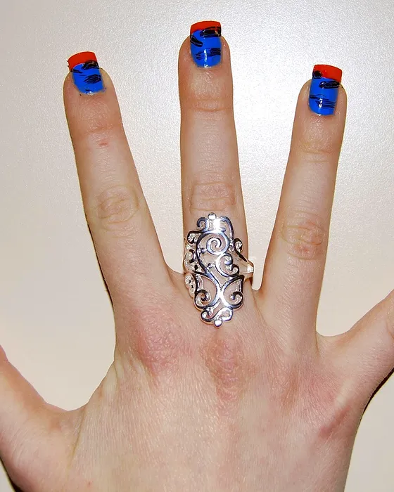 Mão aberta com unhas nude e pontas em azul e vermelho, e um anel de prata.