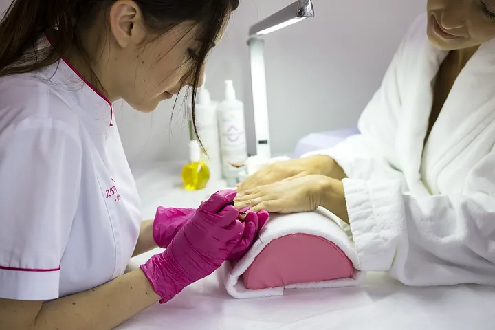 Uma manicure de luvas rosa prepara a mão de uma cliente junto à lâmpada de cura.